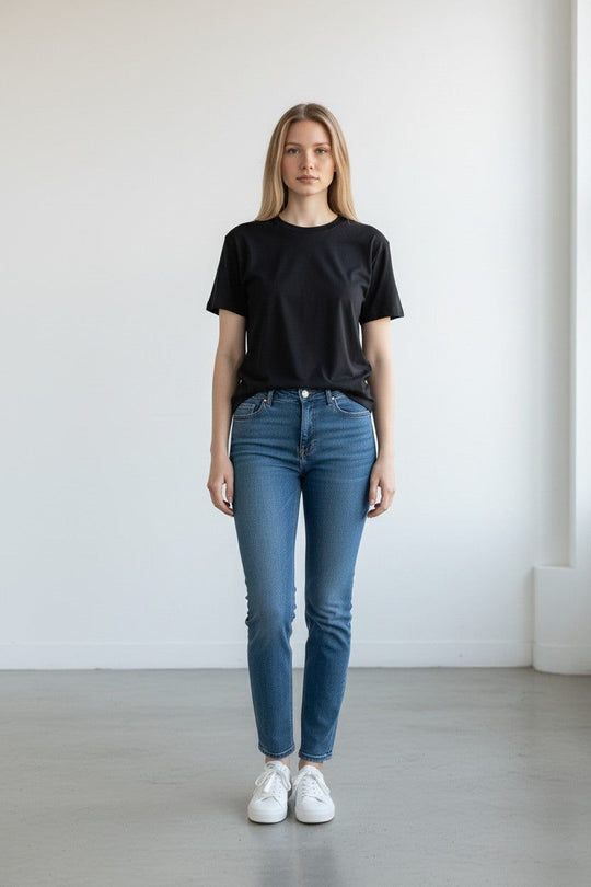 Person wearing a black t-shirt and blue jeans standing against a white wall.