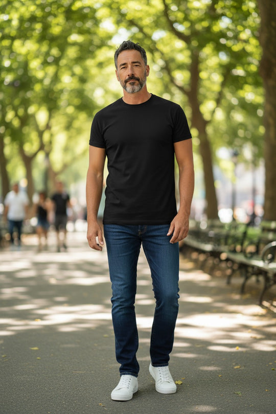 Man wearing a black t-shirt, blue jeans, and white sneakers standing in a park.