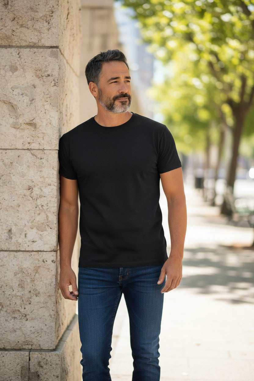 Man wearing a black t-shirt and blue jeans standing against a stone wall with trees in the background.