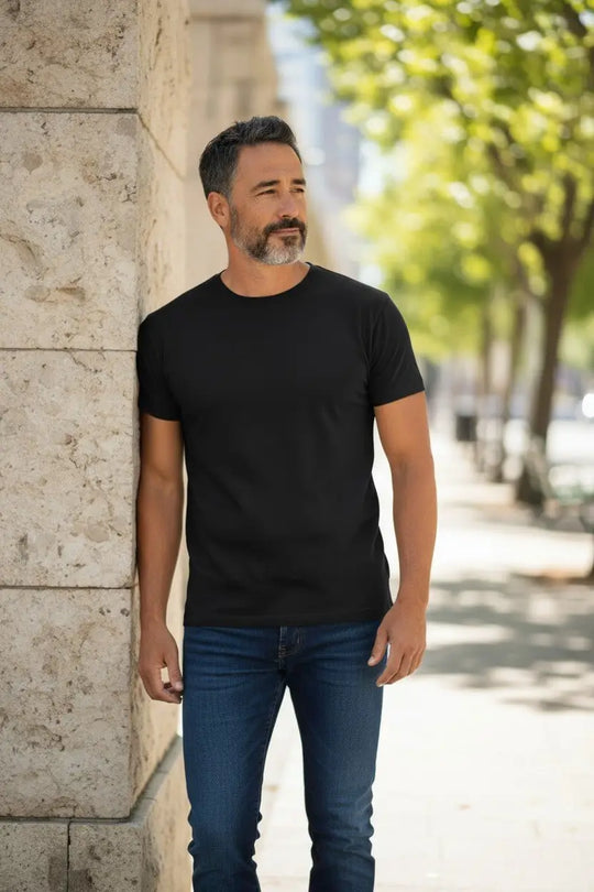 Man wearing a black t-shirt and blue jeans standing against a stone wall with trees in the background.