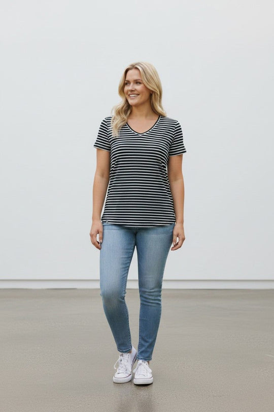 A woman modeling a black and white horizontally striped short-sleeve V-neck t-shirt, paired with light blue skinny jeans and white sneakers, standing against a plain white wall.