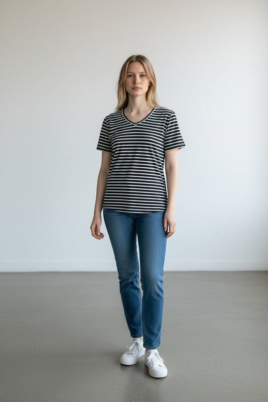 A woman stands in a minimalist studio wearing a black and white horizontally striped V-neck t-shirt, blue jeans, and white sneakers.