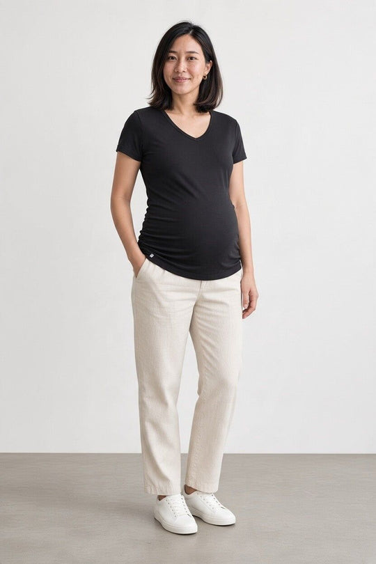 A pregnant woman stands in a studio wearing a black V-neck maternity t-shirt, off-white linen trousers, and white sneakers against a light gray background.