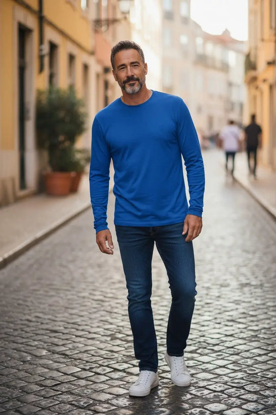 Man wearing a blue long-sleeve shirt and jeans standing on a cobblestone street.