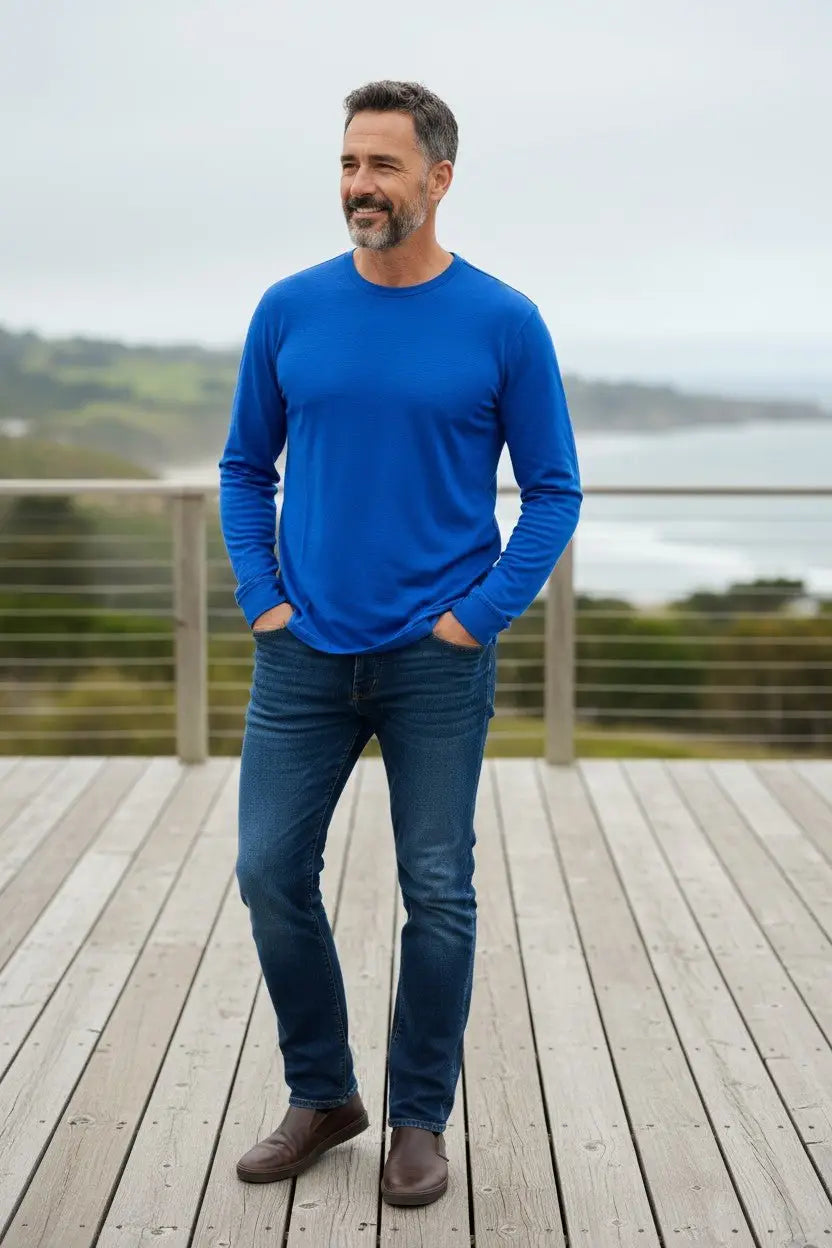 Man wearing a blue long-sleeve shirt and jeans standing on a wooden deck with a scenic background.