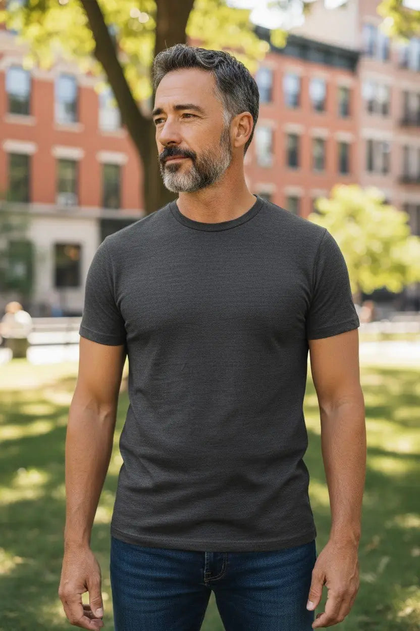 Man wearing a dark gray t-shirt and jeans standing outdoors with buildings and trees in the background