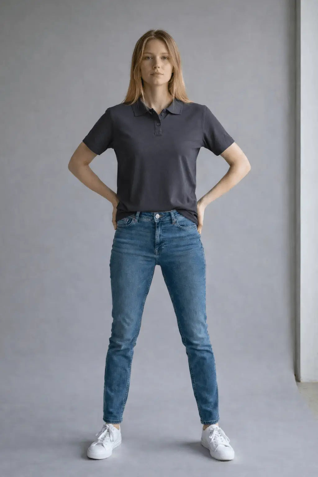 Woman wearing a dark gray polo shirt and blue jeans standing against a plain background