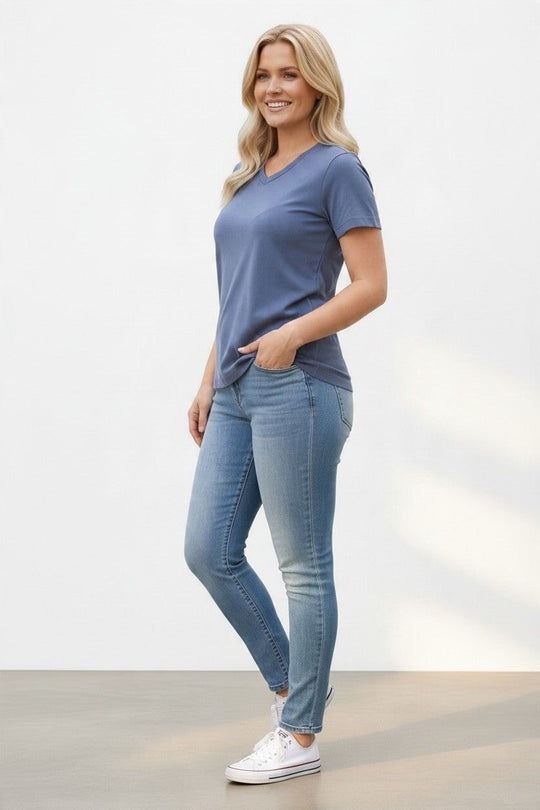 A woman modeling a slate blue v-neck t-shirt and light-wash blue skinny jeans with white sneakers, standing against a clean white studio background.