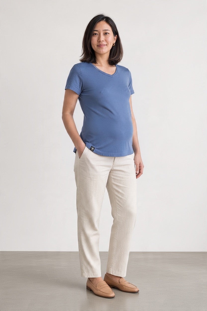 A woman modeling a short-sleeve blue V-neck t-shirt and off-white trousers, standing against a neutral light gray studio background.