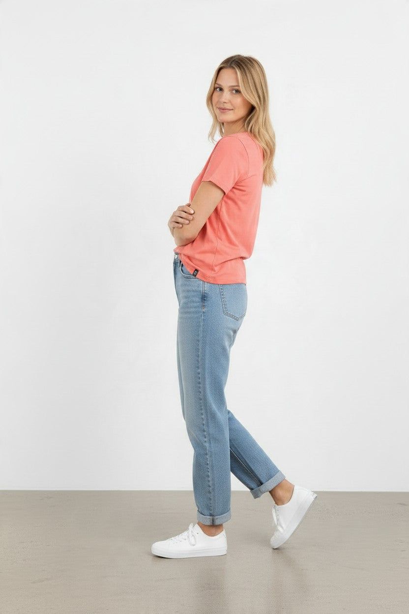 A woman posing in a studio setting, wearing a coral pink t-shirt, light blue cropped jeans, and white sneakers.