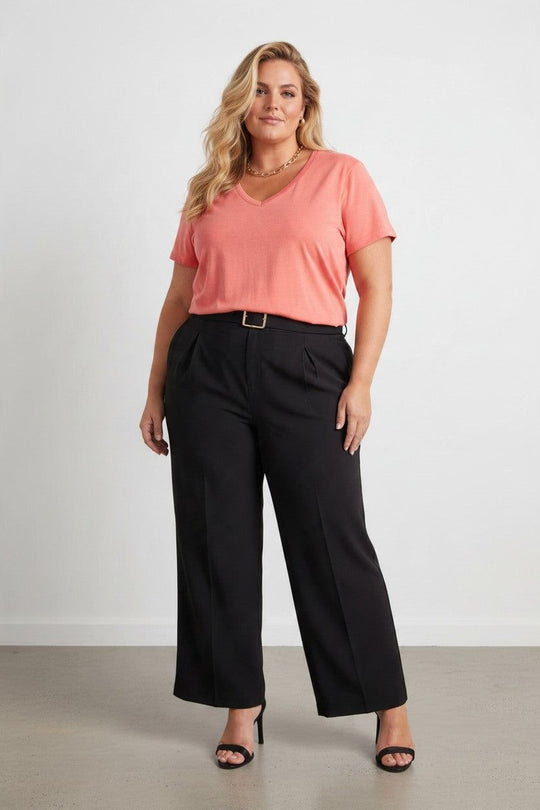 A woman modeling a coral pink V-neck short-sleeve top and black wide-leg trousers with a thin gold-tone belt, standing against a light gray studio wall.