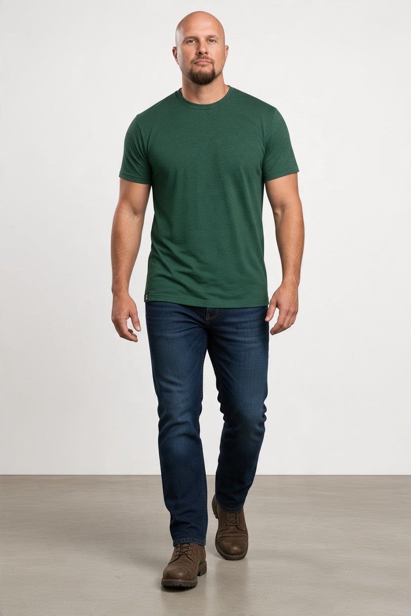 A man wearing a dark green crew-neck t-shirt, dark blue jeans, and brown shoes walking forward against a plain light gray studio background.