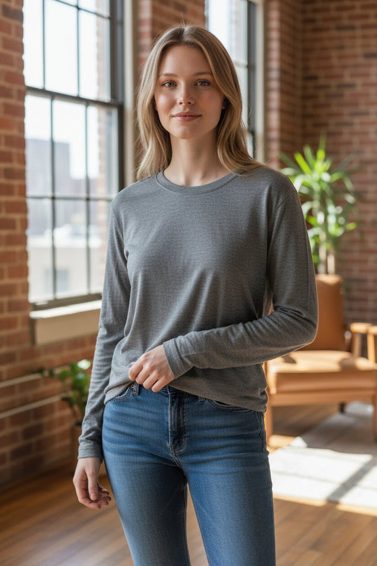 Woman wearing a gray long-sleeve shirt and blue jeans standing in a room with large windows and a plant.