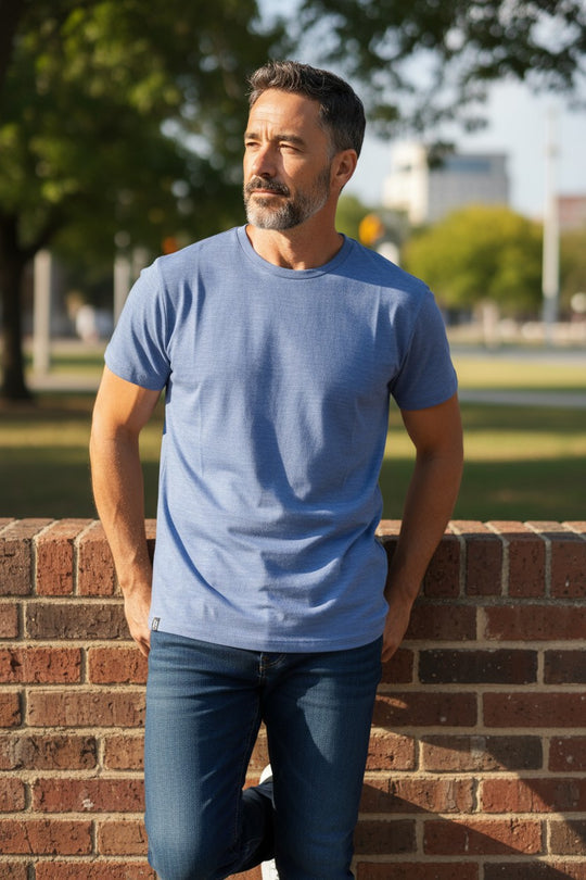 Man wearing a blue t-shirt and jeans standing against a brick wall with trees and buildings in the background.