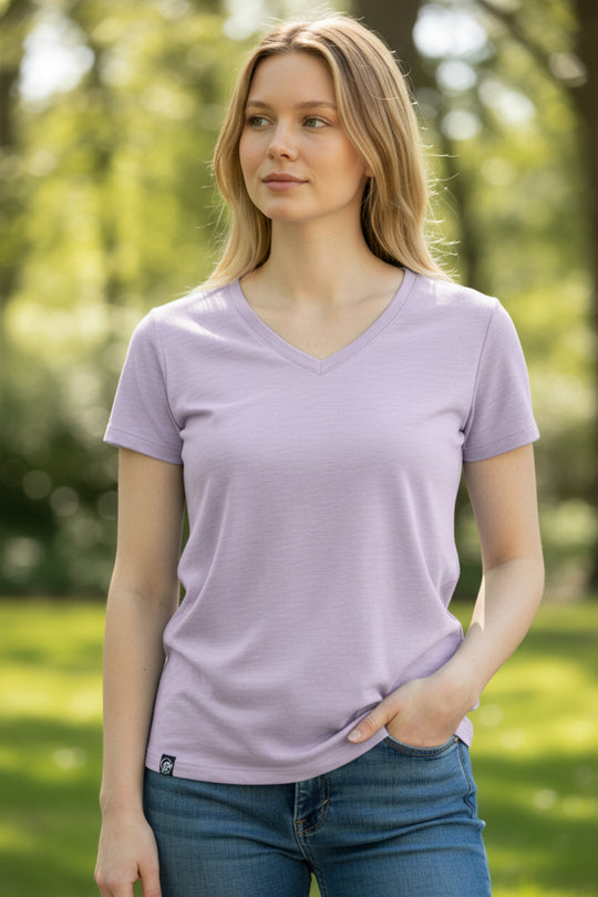 Woman wearing a light purple t-shirt and blue jeans standing outdoors with greenery in the background