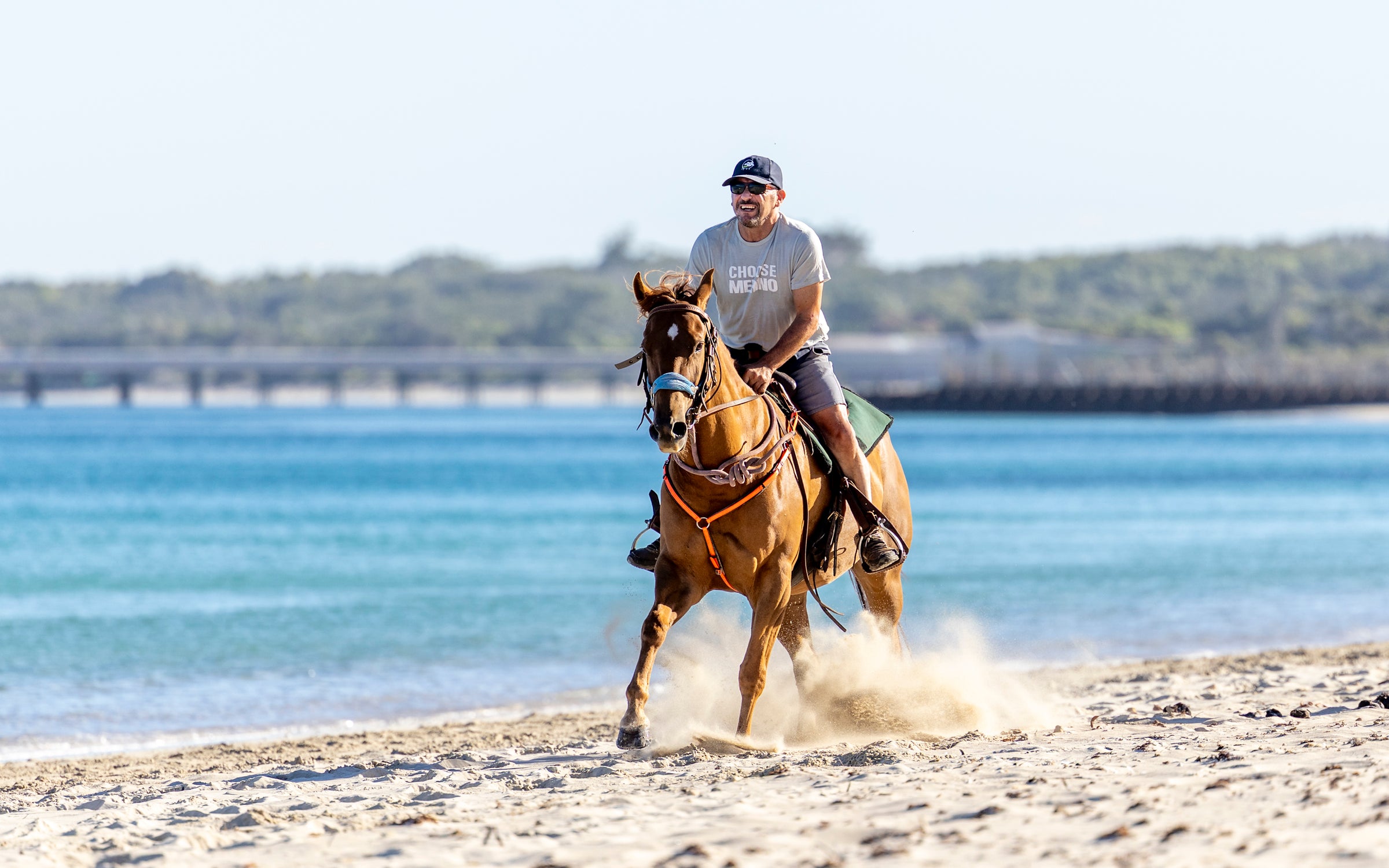 Man riding a horse on a sandy beach with water and a bridge in the background