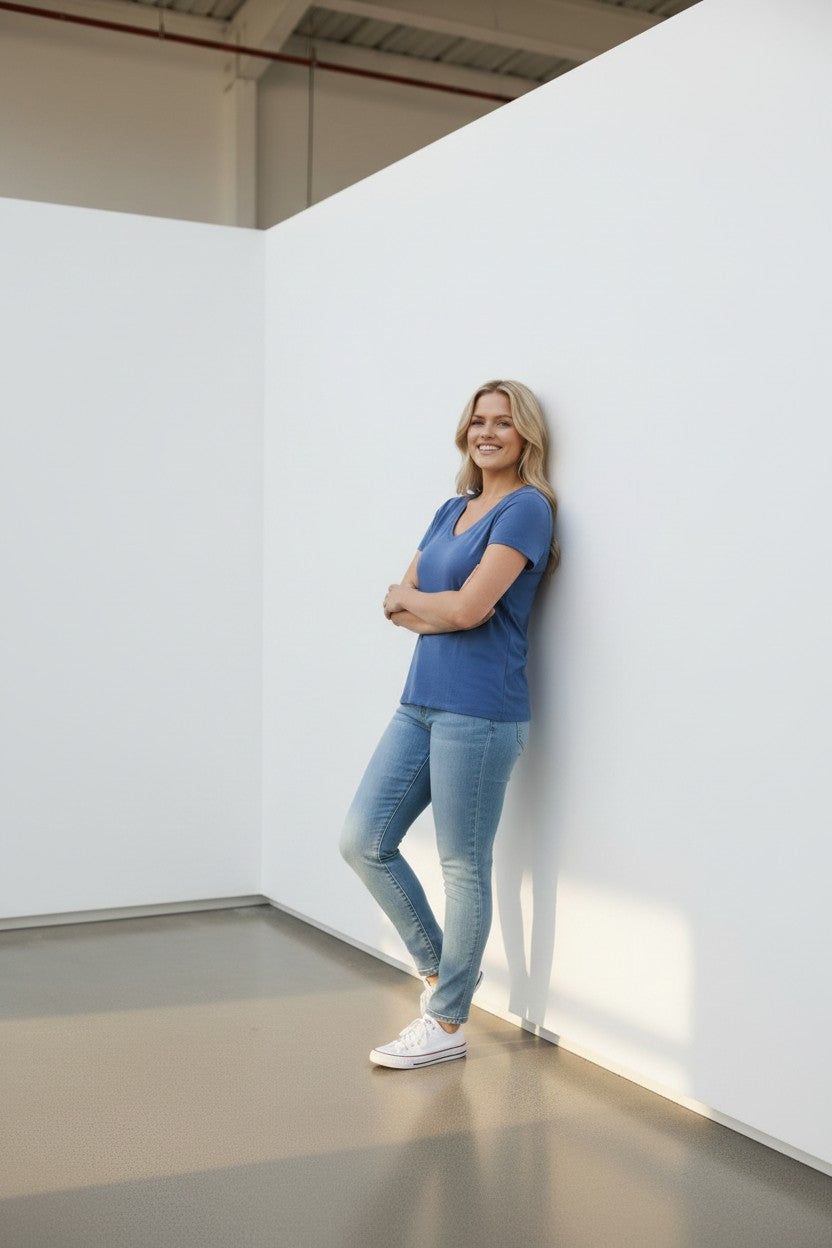 A smiling blonde woman wearing a blue v-neck t-shirt, light-wash blue jeans, and white sneakers leans against a white wall in a minimalist gallery or studio space.