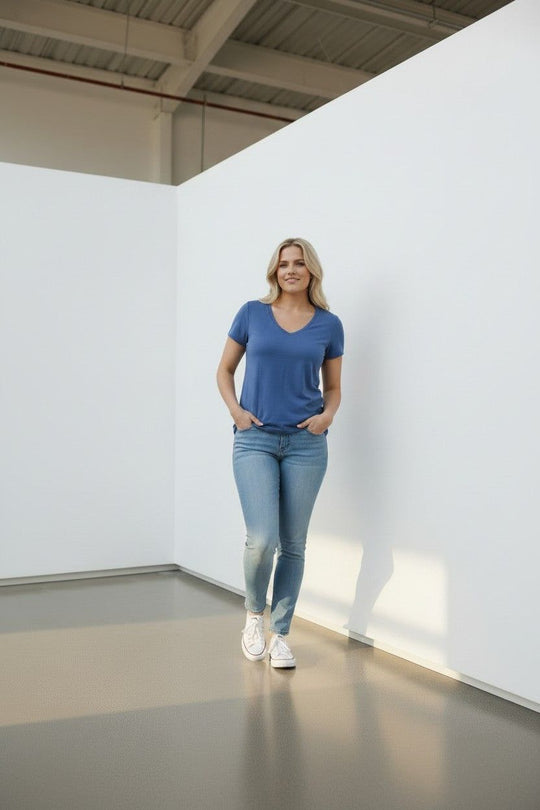 A woman stands in a minimalist white room wearing a blue v-neck t-shirt, light blue skinny jeans, and white sneakers.
