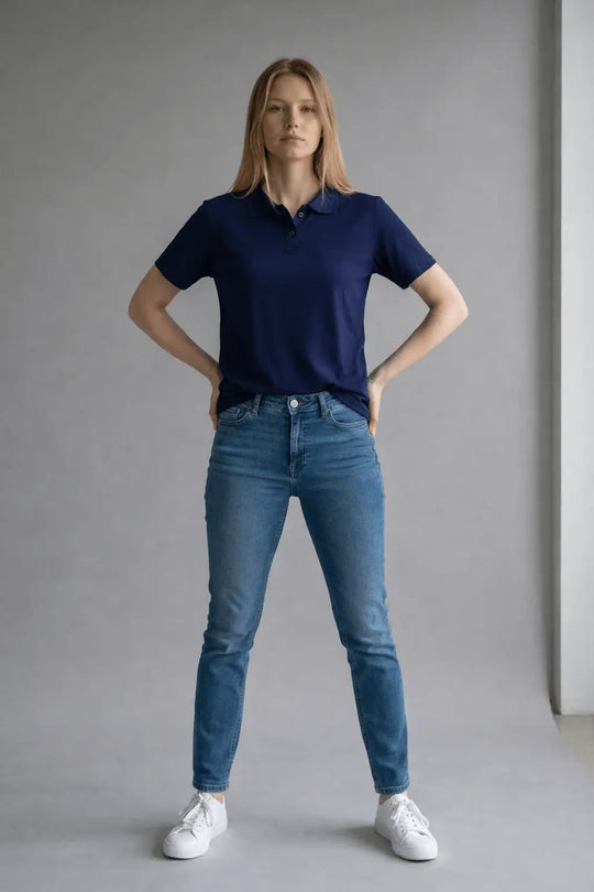 Woman wearing a navy polo shirt and blue jeans standing against a plain background