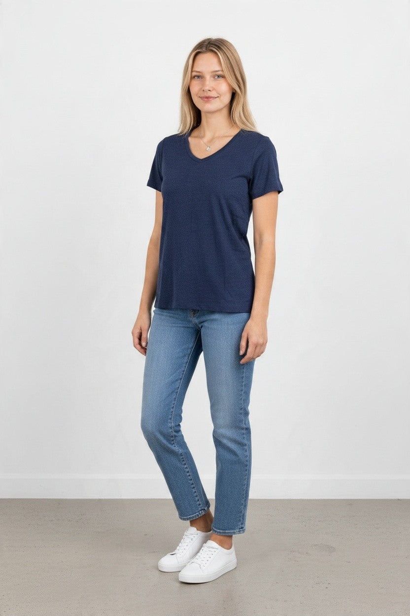 A woman modeling a navy blue v-neck short-sleeve t-shirt, paired with light-wash blue jeans and white sneakers, standing against a plain white wall.