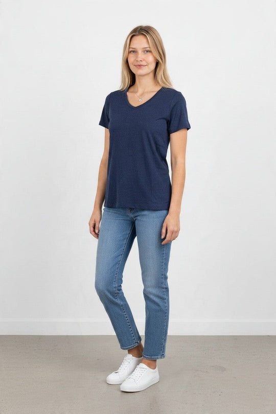 A woman modeling a navy blue v-neck short-sleeve t-shirt, paired with light-wash blue jeans and white sneakers, standing against a plain white wall.