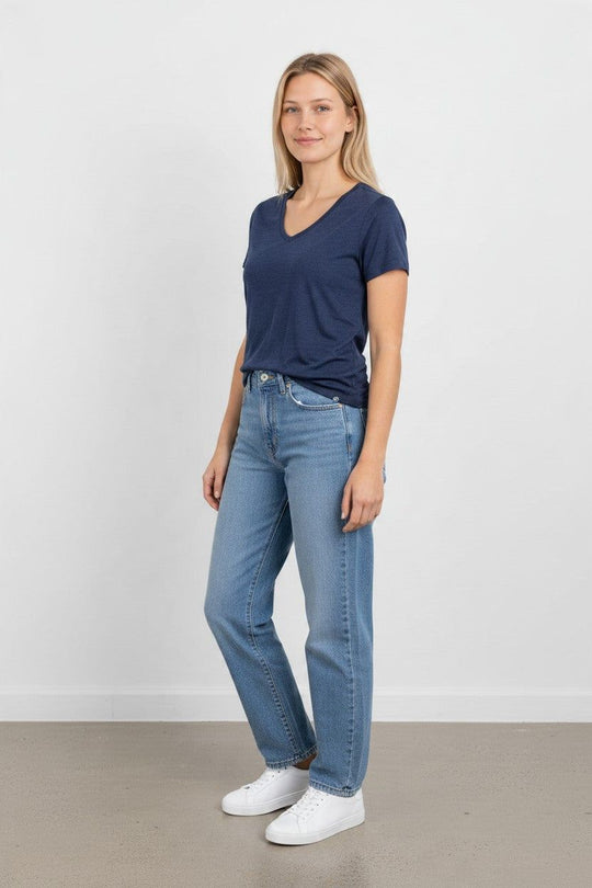 A woman is modeling a navy blue v-neck short-sleeve t-shirt with light-wash blue jeans and white sneakers, posing against a plain white wall.