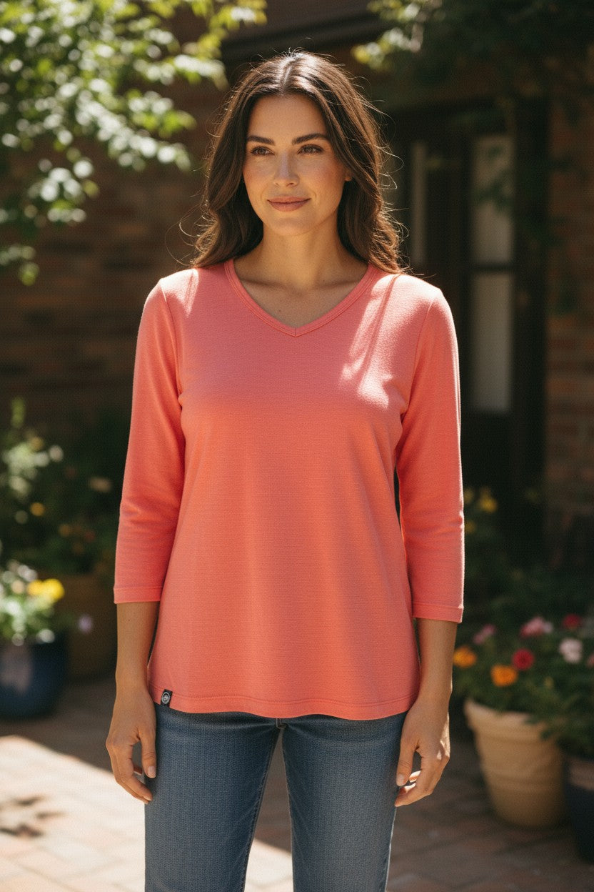 Woman wearing a coral long-sleeve shirt standing outdoors with plants and a building in the background