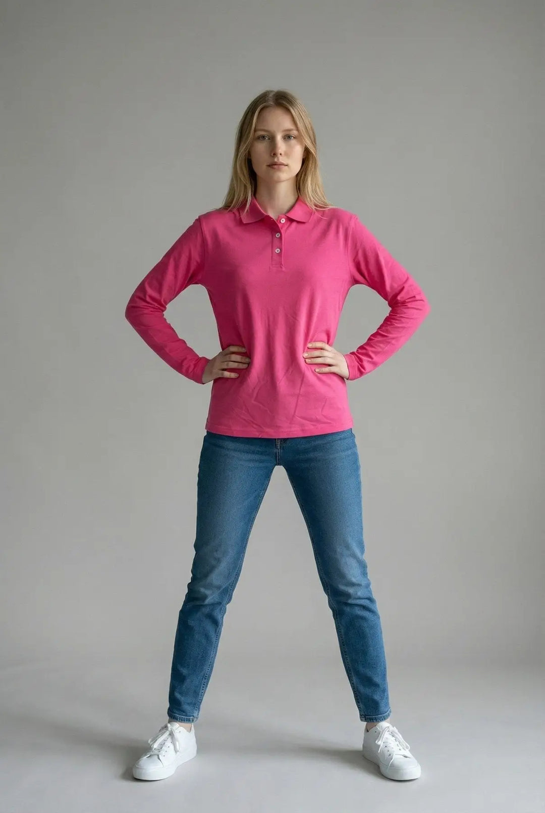 Woman wearing a pink long-sleeve polo shirt and blue jeans against a gray background