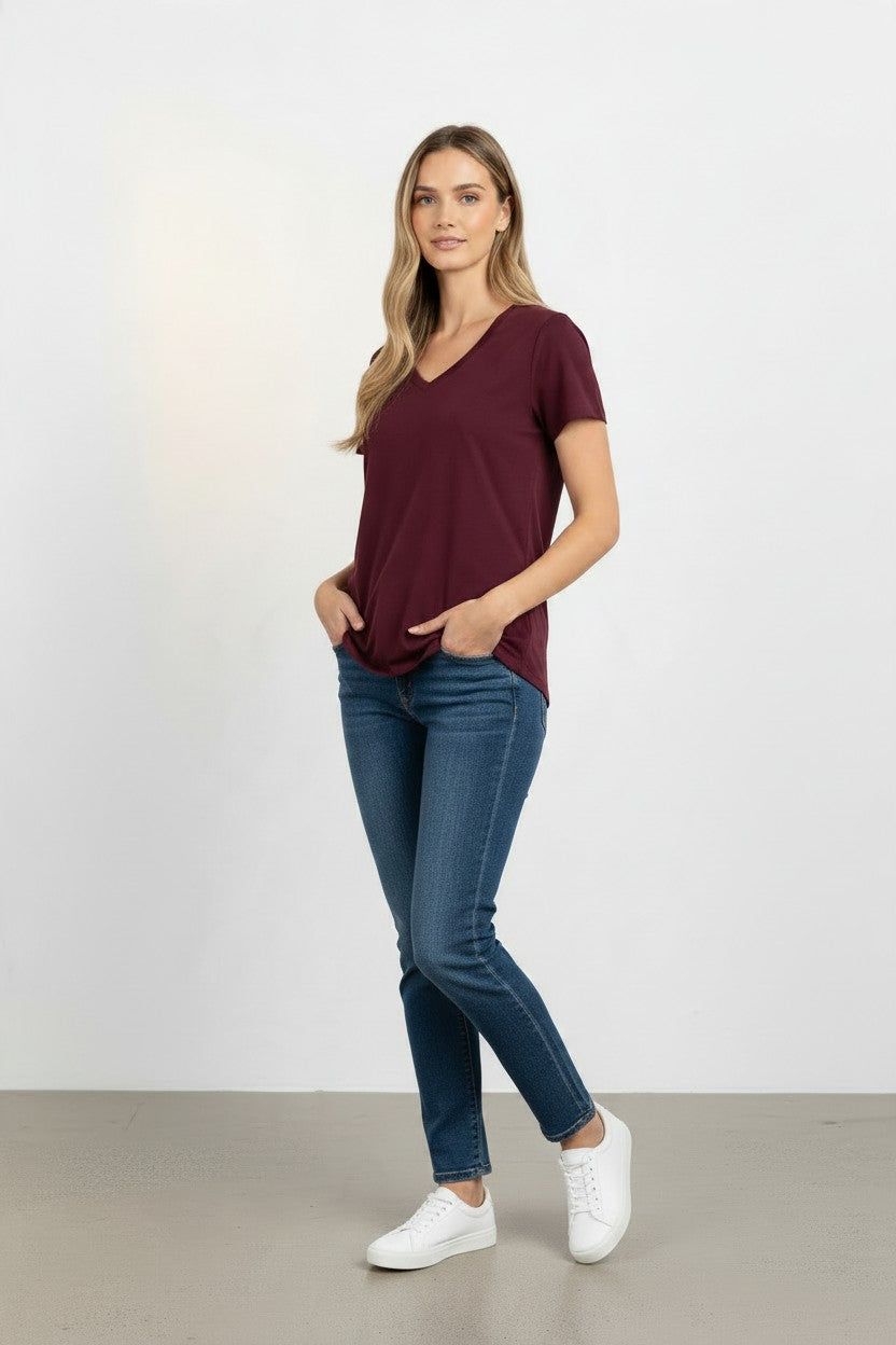 A woman modeling a short-sleeve v-neck t-shirt in burgundy, paired with dark blue skinny jeans and white sneakers, standing against a light gray studio background.