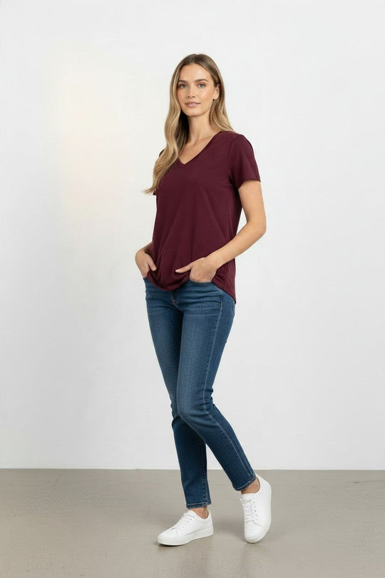 A woman modeling a short-sleeve v-neck t-shirt in burgundy, paired with dark blue skinny jeans and white sneakers, standing against a light gray studio background.