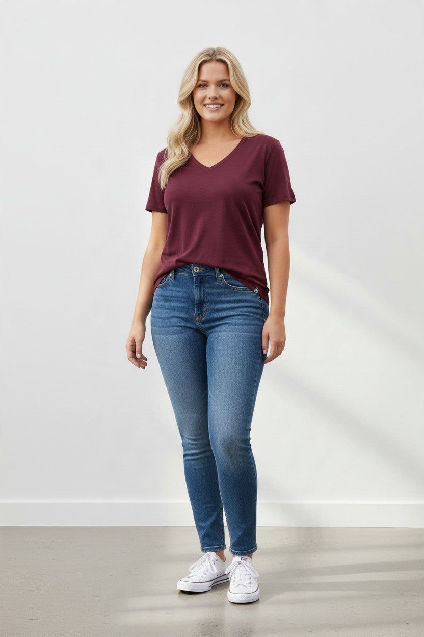A woman modeling a short-sleeve burgundy V-neck t-shirt and blue skinny jeans with white sneakers, standing against a light gray wall.