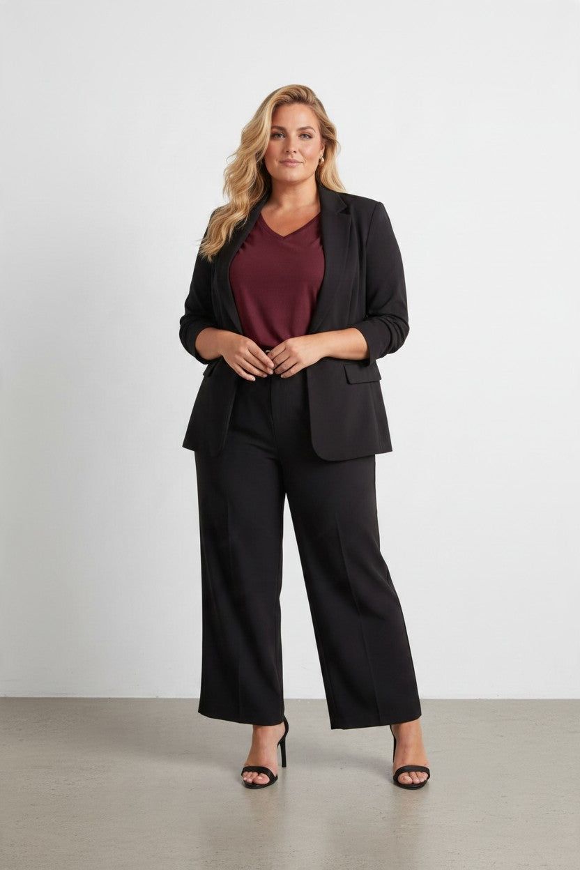 A woman stands in a studio wearing a black tailored blazer and matching wide-leg pants over a burgundy V-neck top, with black heeled sandals.