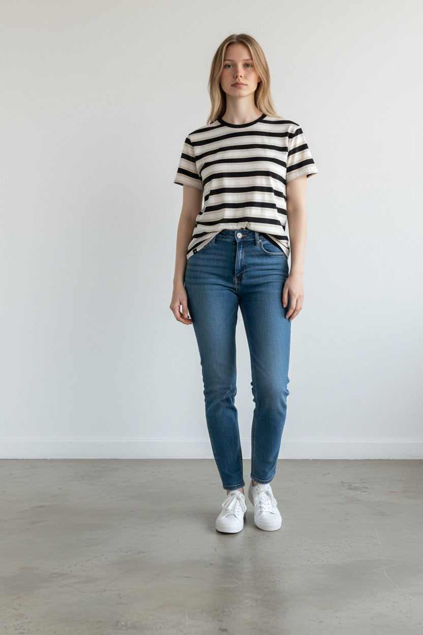 A woman stands in a minimalist studio setting wearing a black and white horizontally striped short-sleeve t-shirt, blue skinny jeans, and white sneakers.