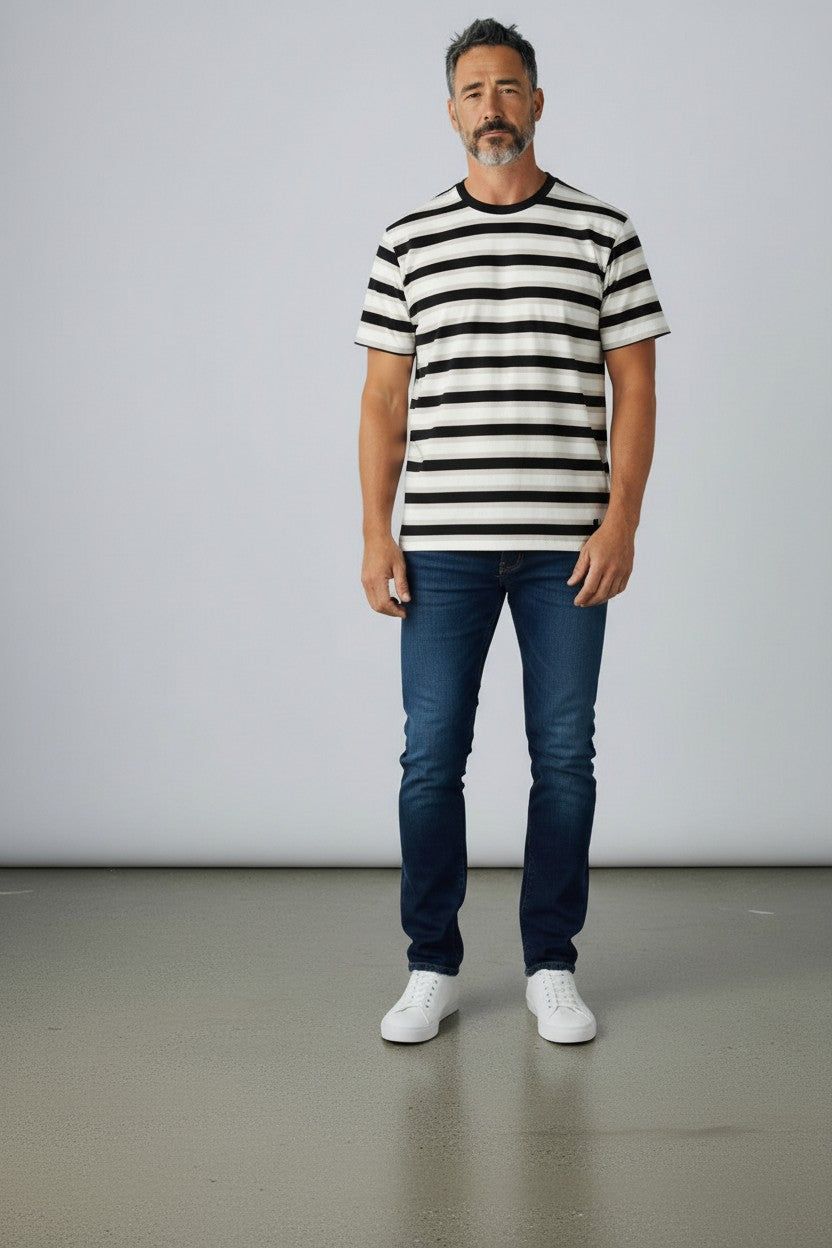 A man wearing a black and white horizontally striped short-sleeve t-shirt, dark blue jeans, and white sneakers, standing against a light gray studio background.