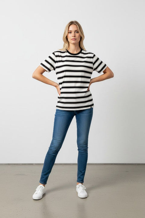 A woman modeling a black and white horizontally striped short-sleeve t-shirt, paired with blue skinny jeans and white sneakers, standing against a plain light gray studio background.