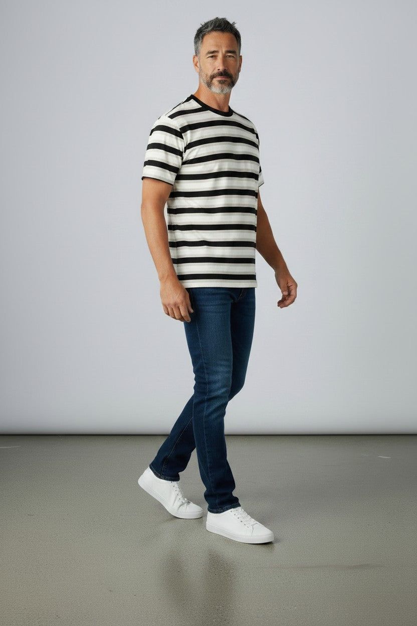 A man wearing a black and white horizontally striped short-sleeve t-shirt, dark blue jeans, and white sneakers, standing against a light gray studio background.