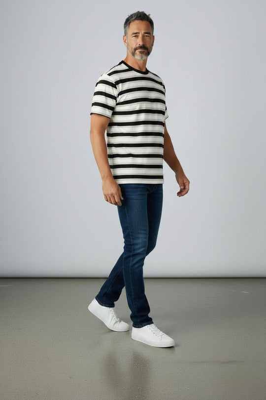 A man wearing a black and white horizontally striped short-sleeve t-shirt, dark blue jeans, and white sneakers, standing against a light gray studio background.