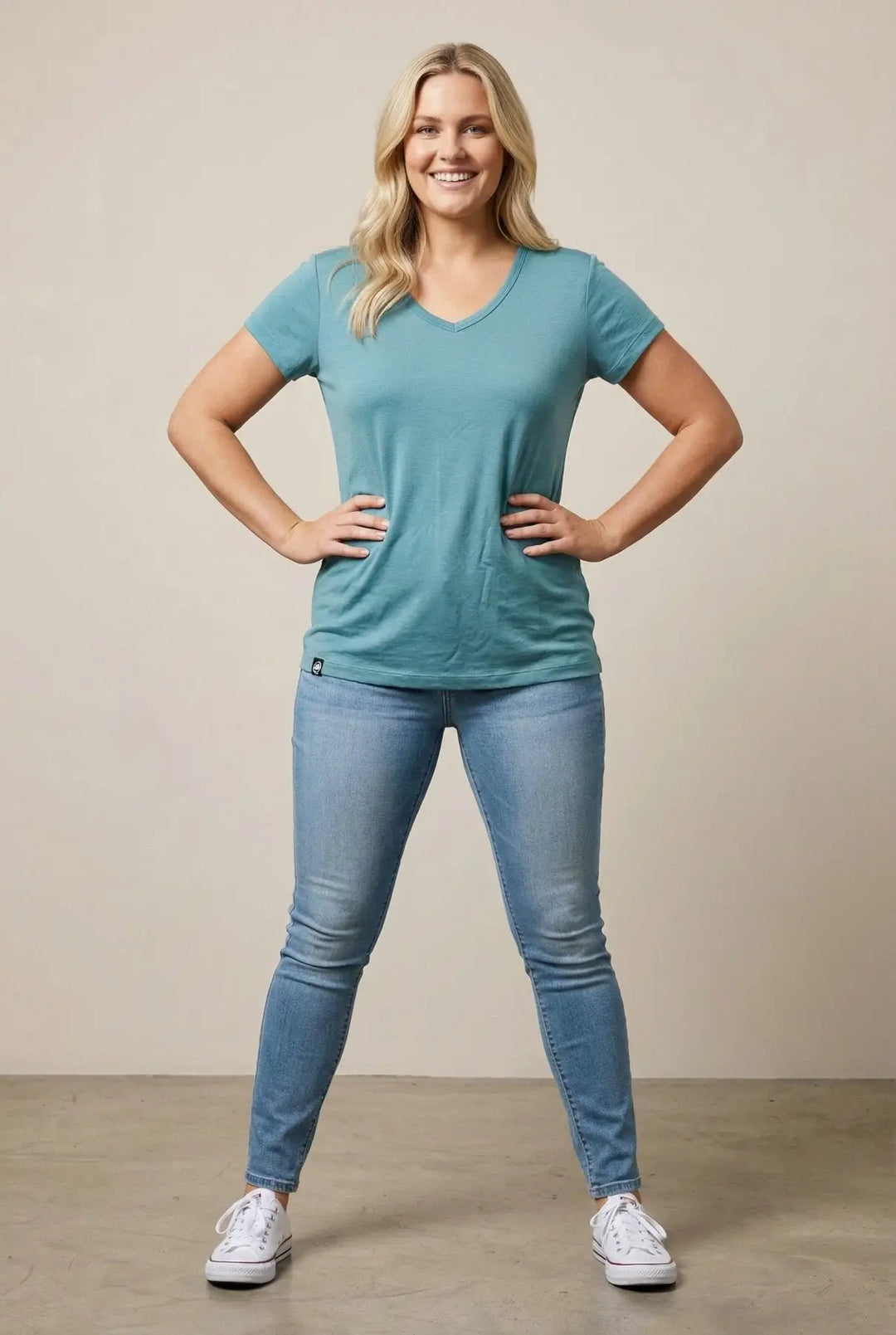 Woman wearing a teal t-shirt and blue jeans standing against a beige background