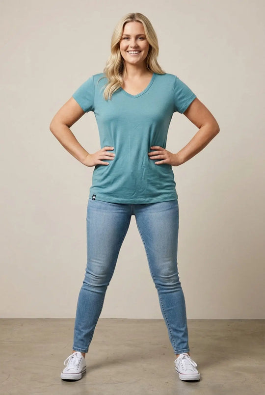 Woman wearing a teal t-shirt and blue jeans standing against a beige background