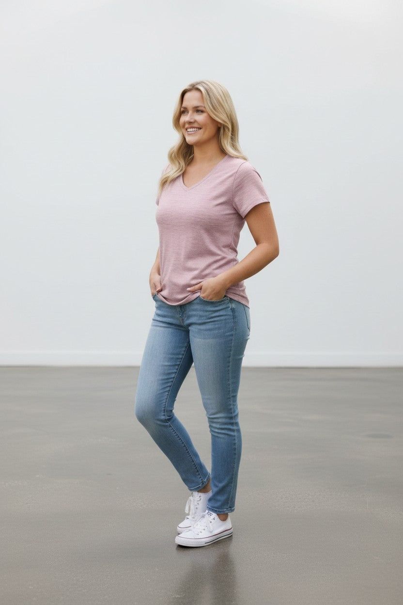 A woman modeling a light pink v-neck t-shirt, blue skinny jeans, and white sneakers in a minimalist studio setting with a white wall and gray floor.