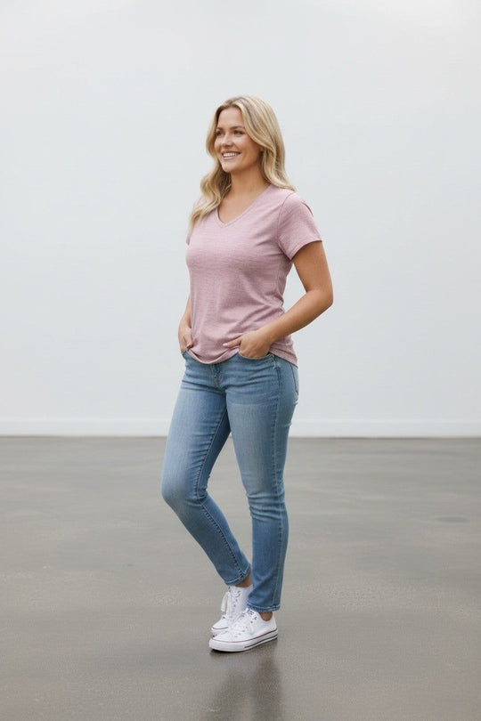 A woman modeling a light pink v-neck t-shirt, blue skinny jeans, and white sneakers in a minimalist studio setting with a white wall and gray floor.