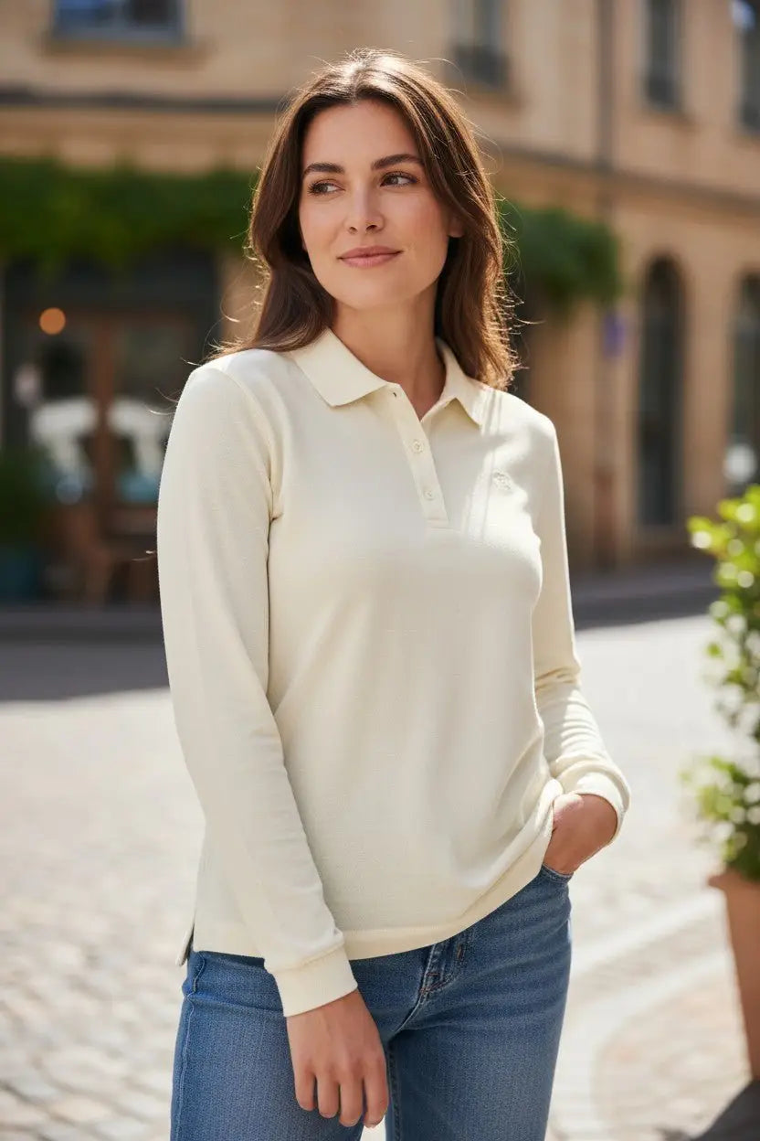 Woman wearing a cream long-sleeve polo shirt and blue jeans standing on a street.