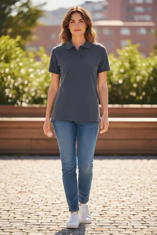 Woman wearing a dark gray polo shirt and blue jeans standing on a paved area with greenery and buildings in the background.