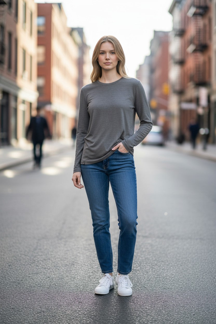 Woman wearing a gray long-sleeve top and blue jeans standing on a city street.