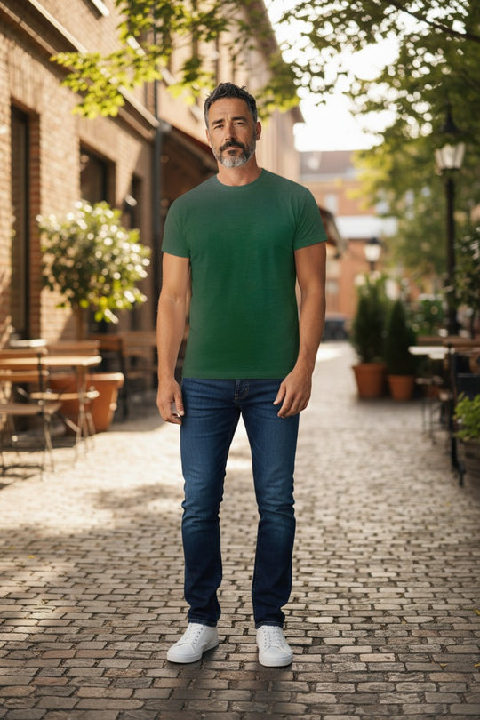 Man wearing a green t-shirt and blue jeans standing on a cobbled street with trees and buildings in the background.