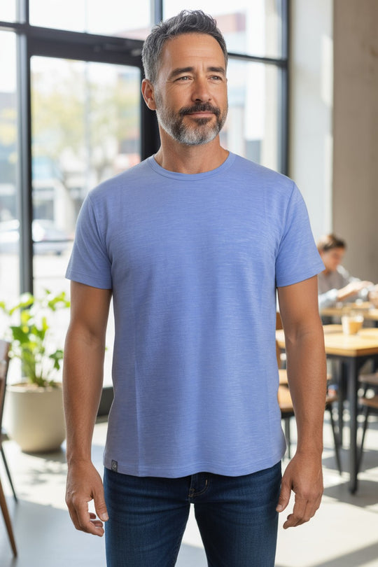 Man wearing a blue merino t-shirt standing in a modern indoor setting with large windows.