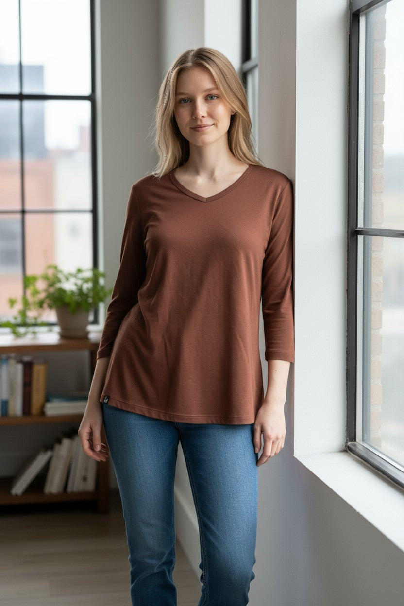Woman wearing a brown top and blue jeans standing in a room with large windows.