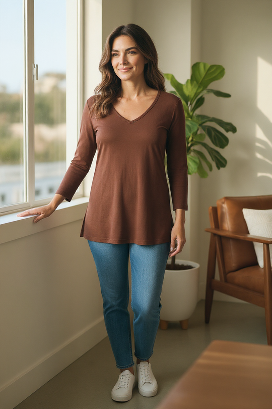 Woman in a brown tunic and blue jeans standing by a window in a bright room.