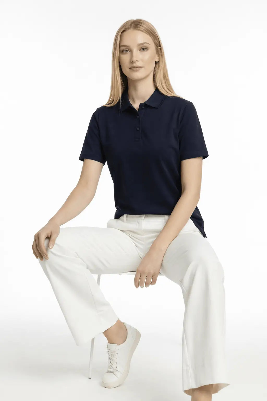 Woman wearing a navy polo shirt and white pants sitting on a white background