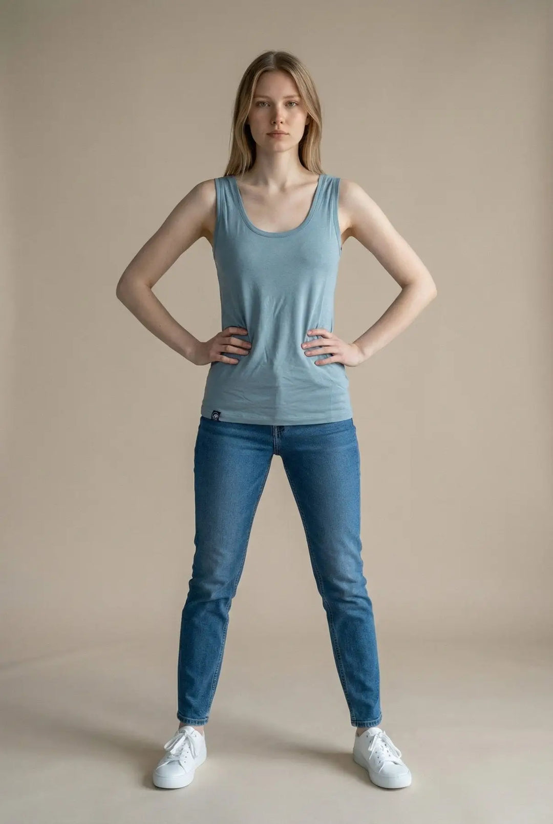 Woman wearing a light blue tank top and jeans standing against a beige background
